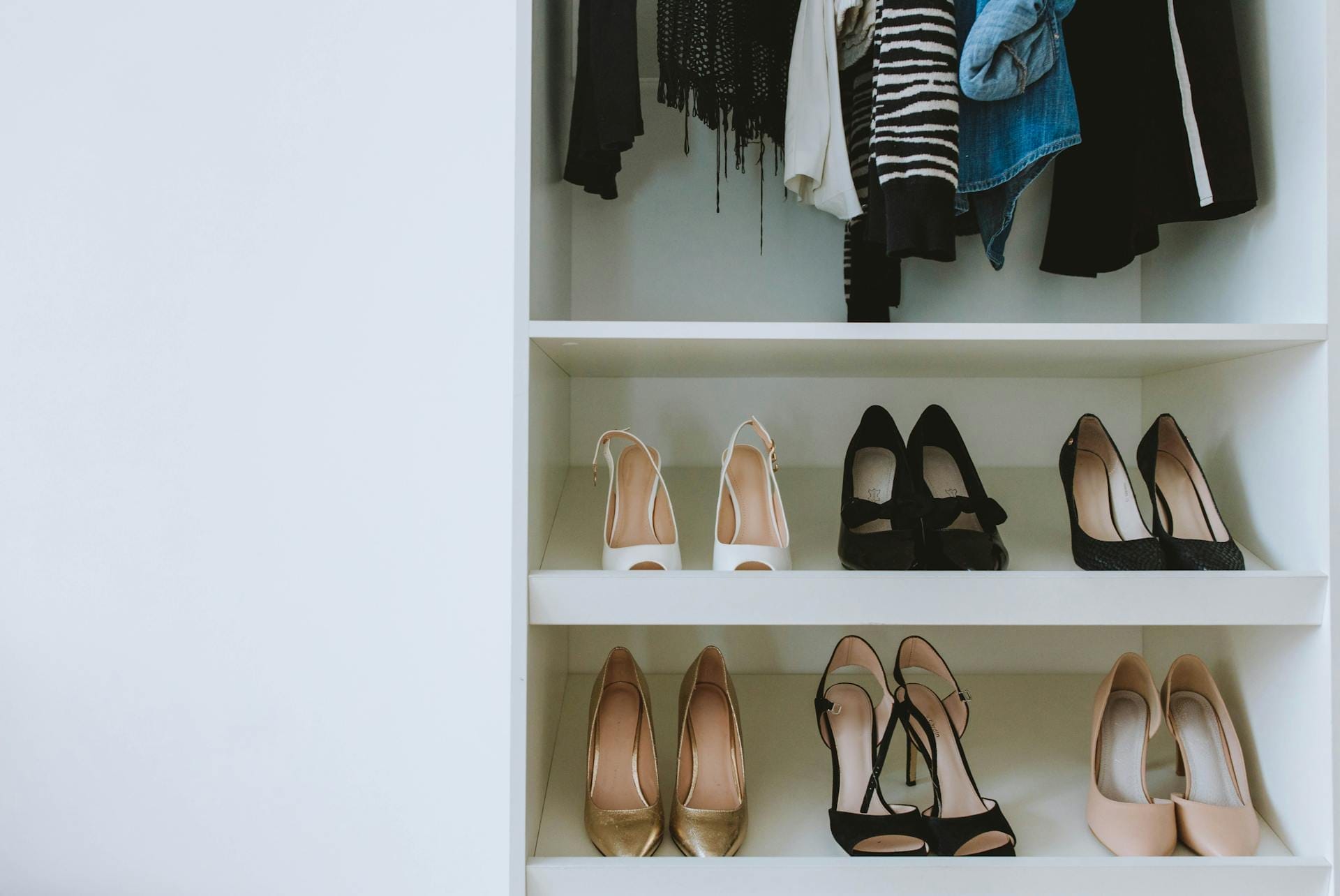 A closet with hanging clothes above three rows of high-heeled shoes in neutral colors, including black and beige, neatly organized on white shelves.