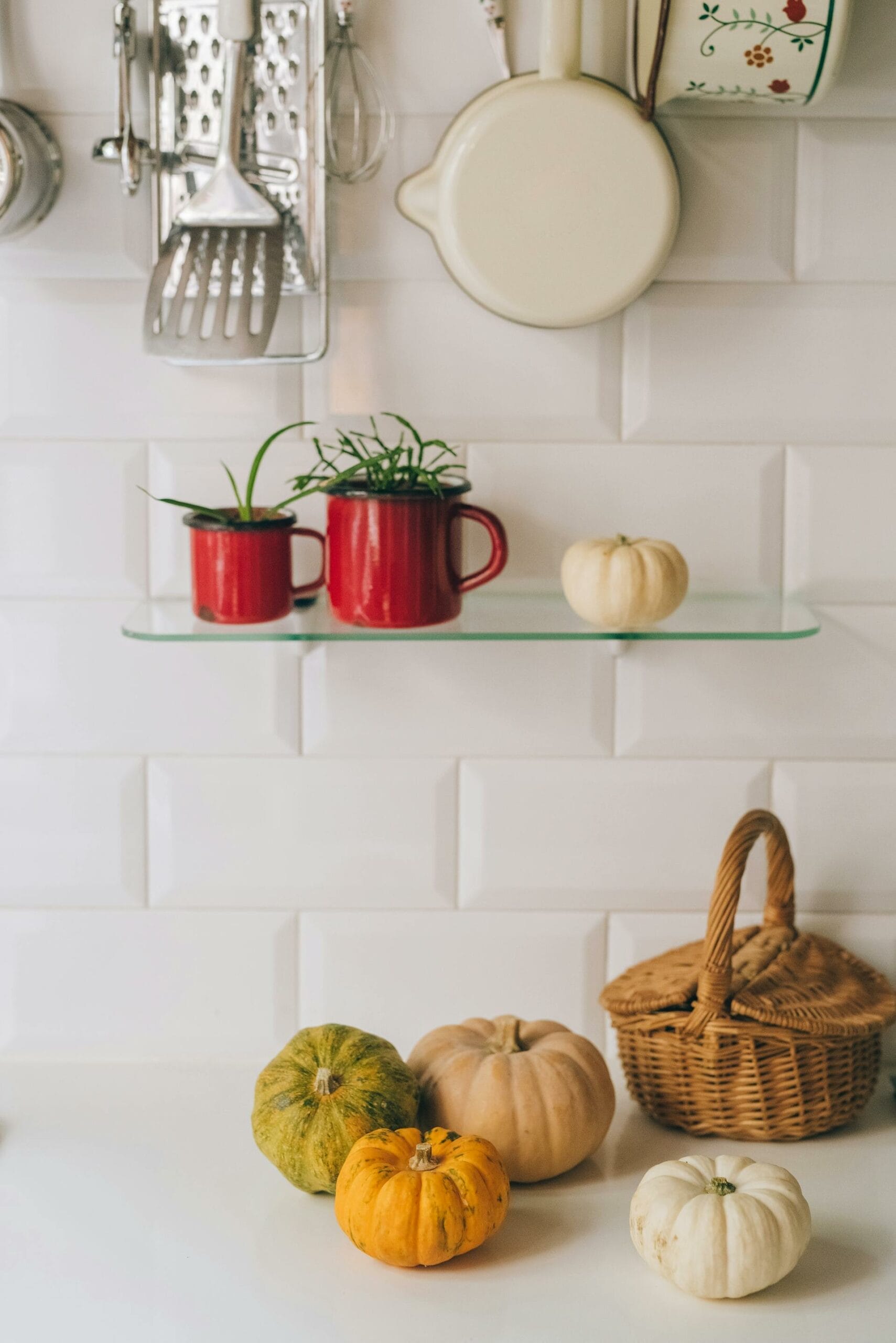 Kitchen counter with three small pumpkins and a wicker basket. Two red mugs with plants are on a glass shelf above, surrounded by hanging kitchen utensils and a pan.