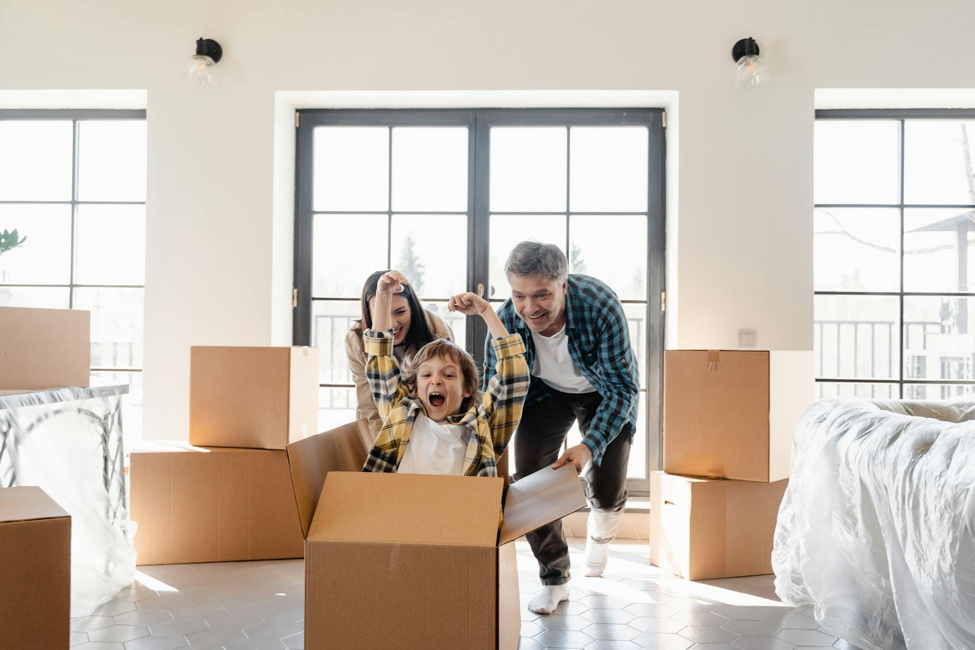 A man and woman play with a child in a cardboard box inside a room with unpacked boxes.