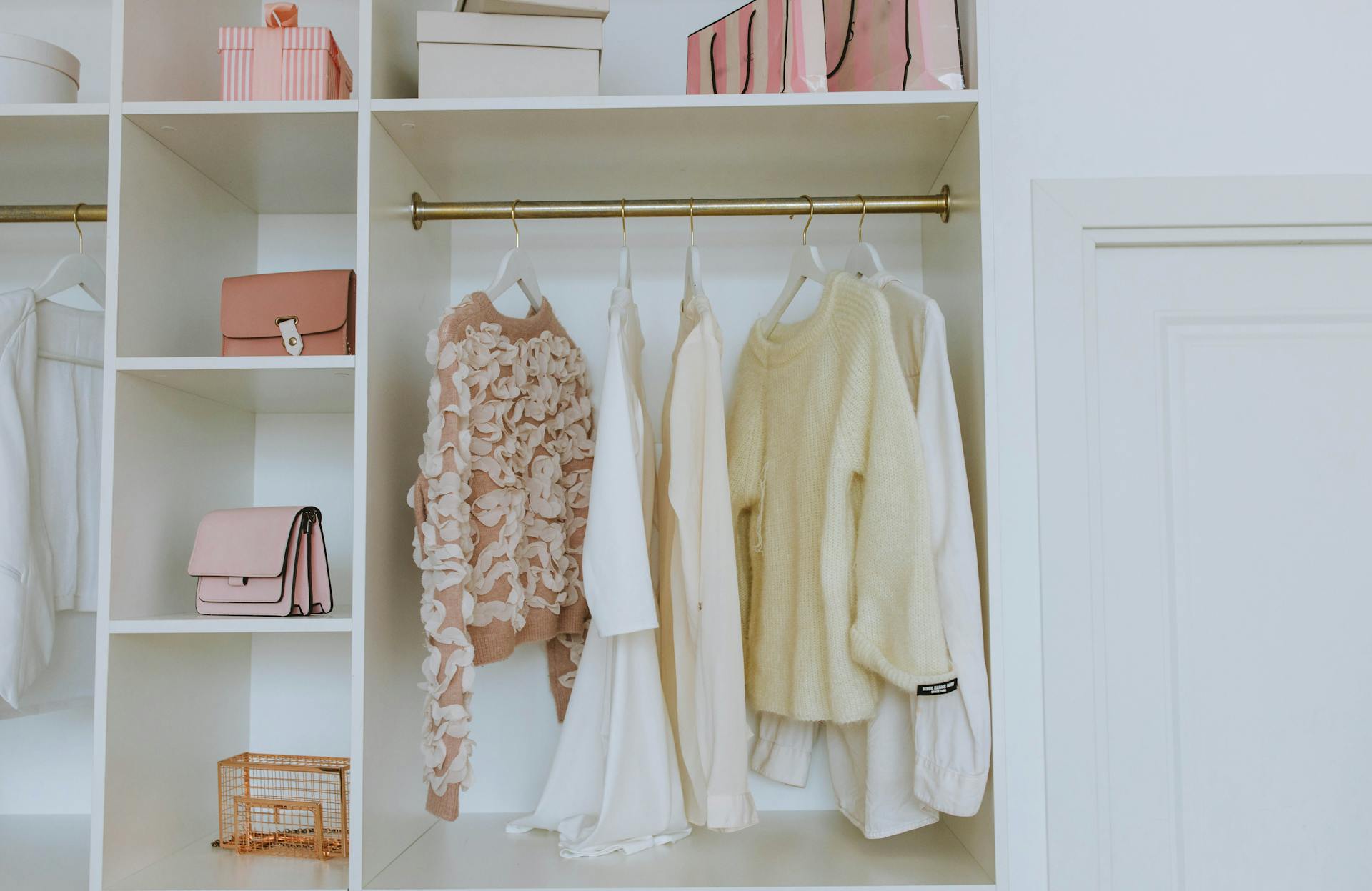 Open closet with a brass rod holding pastel sweaters and blouses on hangers. Shelves contain pink and white boxes, a pink handbag, and a wire basket.