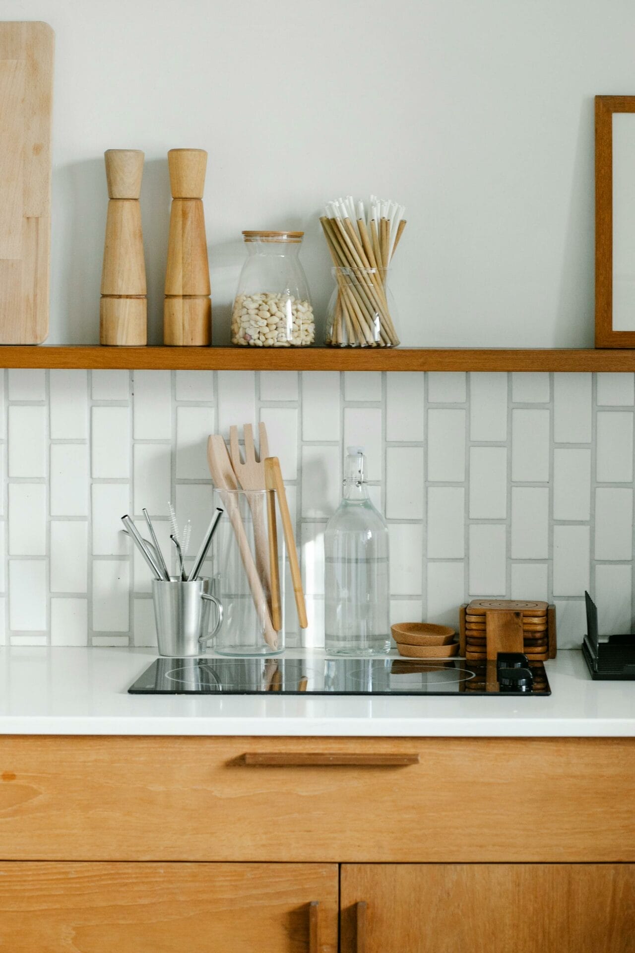 Modern kitchen with wooden cabinets, white tile backsplash, and items like cutting boards, utensils, pepper mills, and a glass jar on the countertop. A glass bottle and a metal utensil holder are also present.