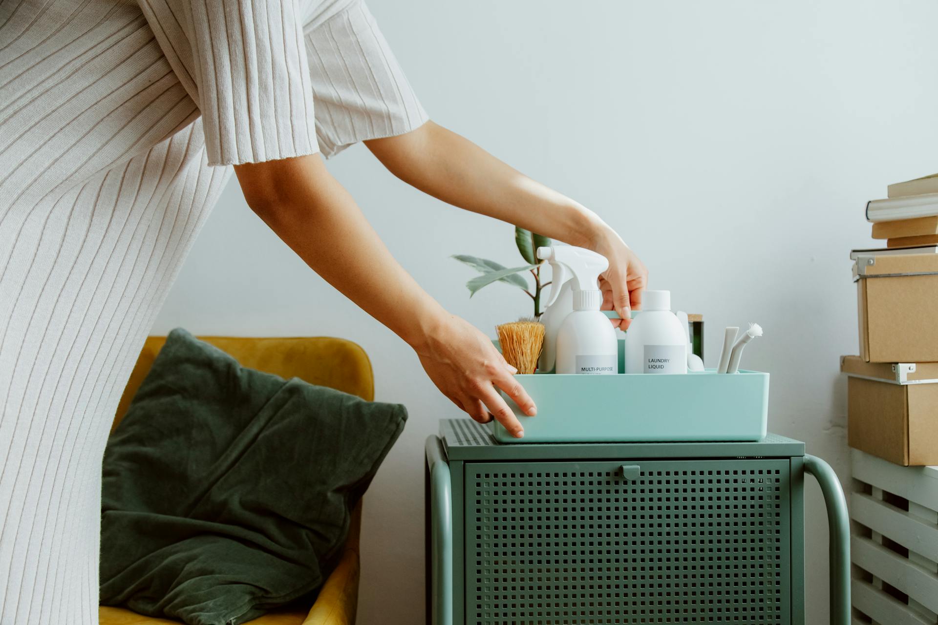 Person organizing a tray with cleaning supplies and a potted plant on a green cabinet next to a yellow armchair.