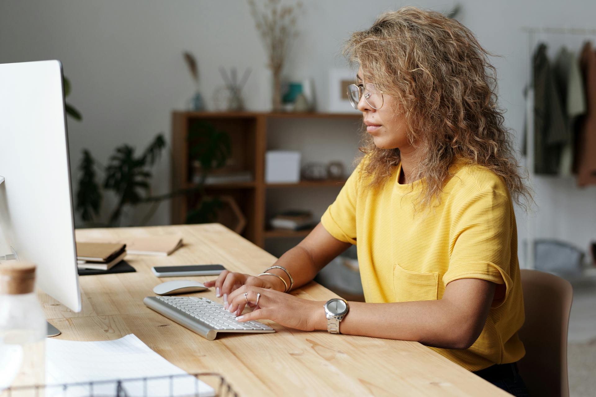 Person in a yellow shirt typing at a desk with a computer in a home office setting.