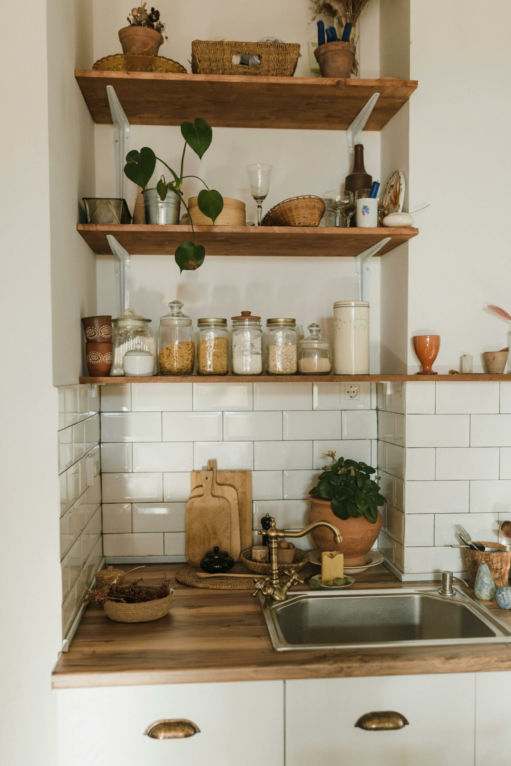 A tidy kitchen corner with open wooden shelves holding plants, jars, and baskets. A sink is below, with a small potted plant and wooden cutting boards on the counter.