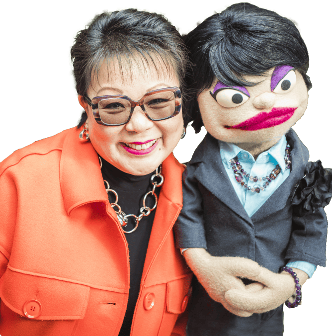 Linda Chu is in an orange coat smiling next to a puppet with exaggerated features and dark hair, wearing a dark blazer.