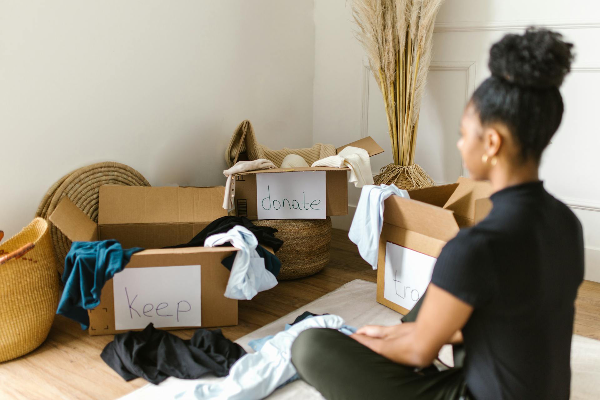 A person sits on the floor facing three labeled boxes: "keep," "donate," and "trash." Clothing items are scattered around and in the boxes.