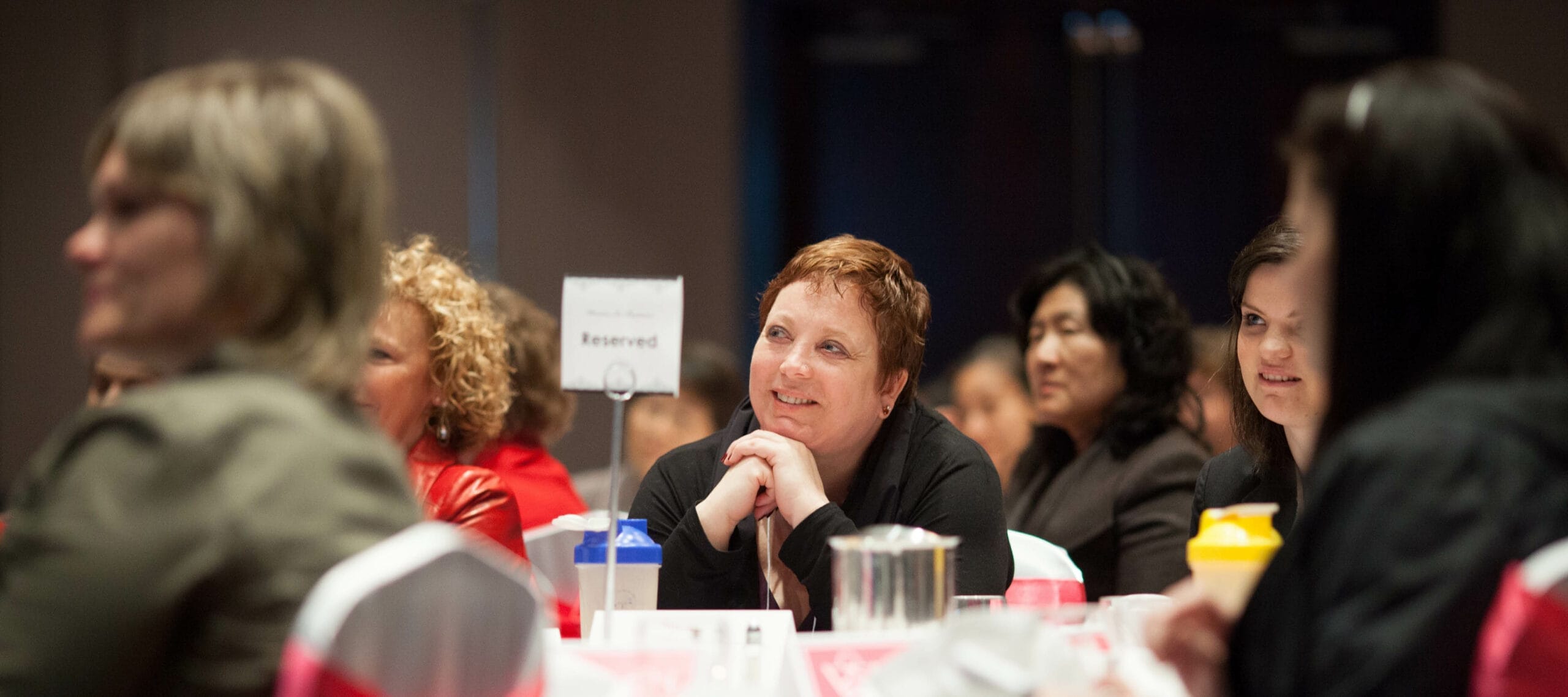 A group of people seated at a round table during an event, with a reserved sign in the center. A woman in the foreground is smiling attentively with her hands resting on her chin, surrounded by others engaged in the event.