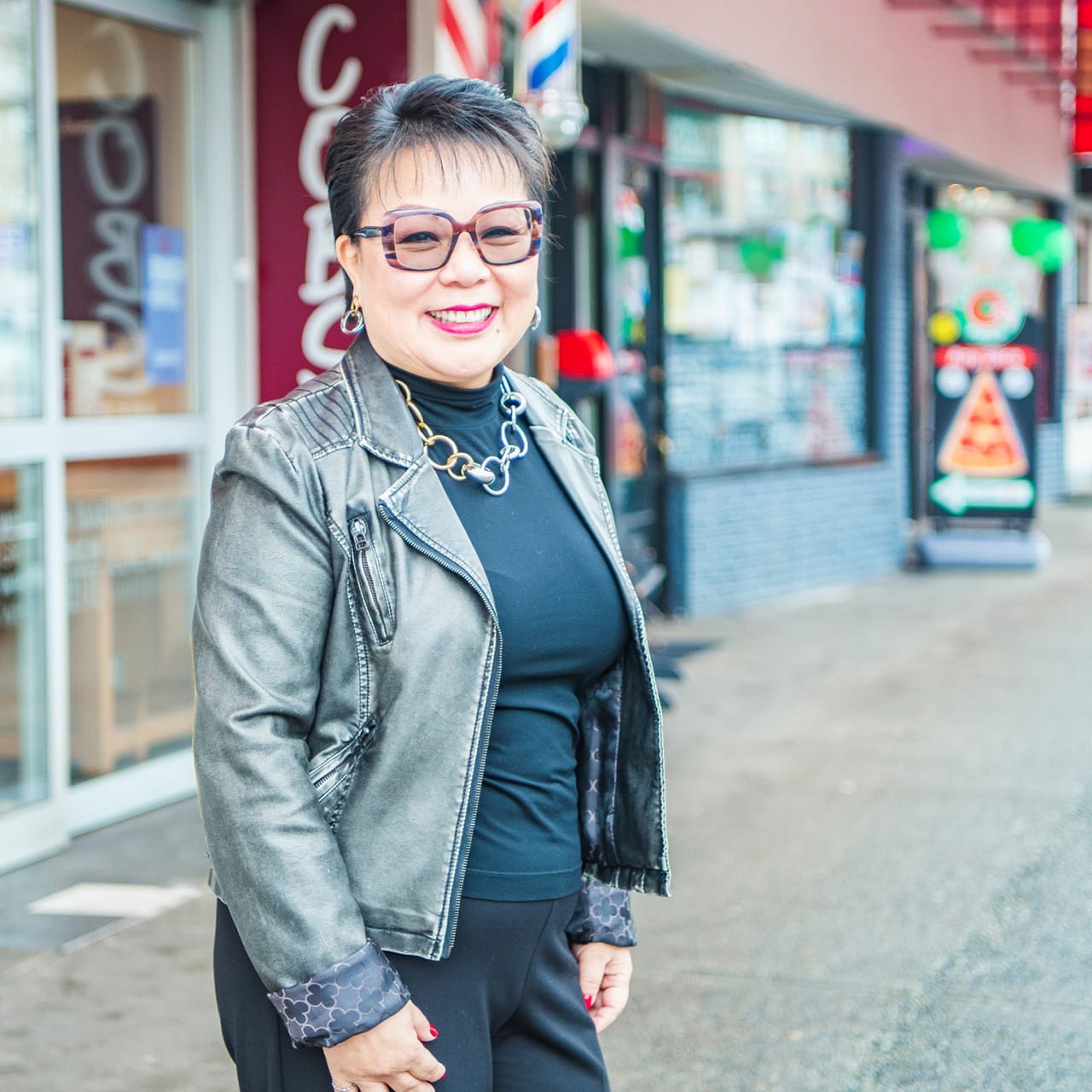 Linda Chu is wearing a leather jacket and glasses. She stands smiling on a city street.