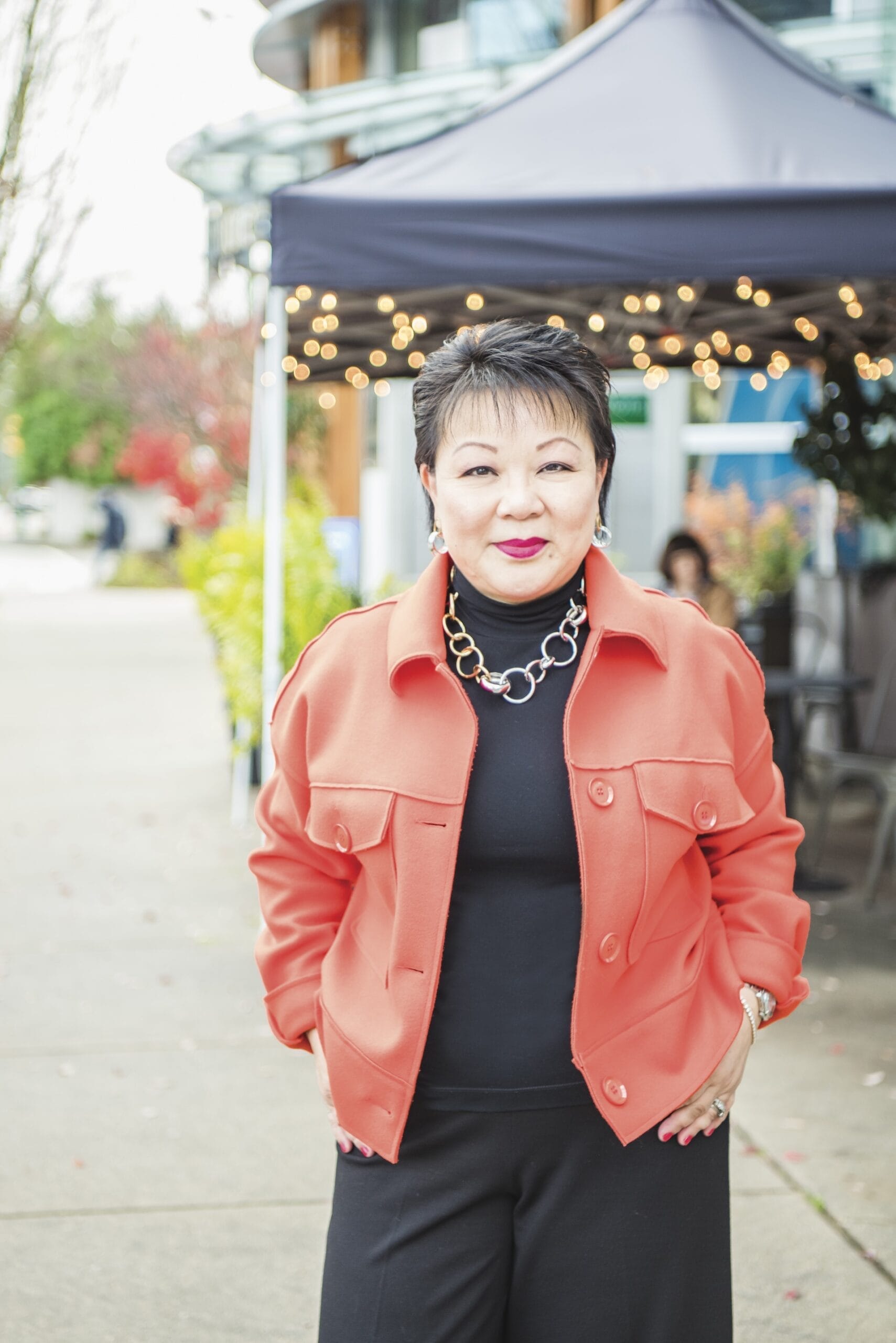 Linda Chu is wearing a coral jacket and black turtleneck, and stands outdoors near a tent with string lights.