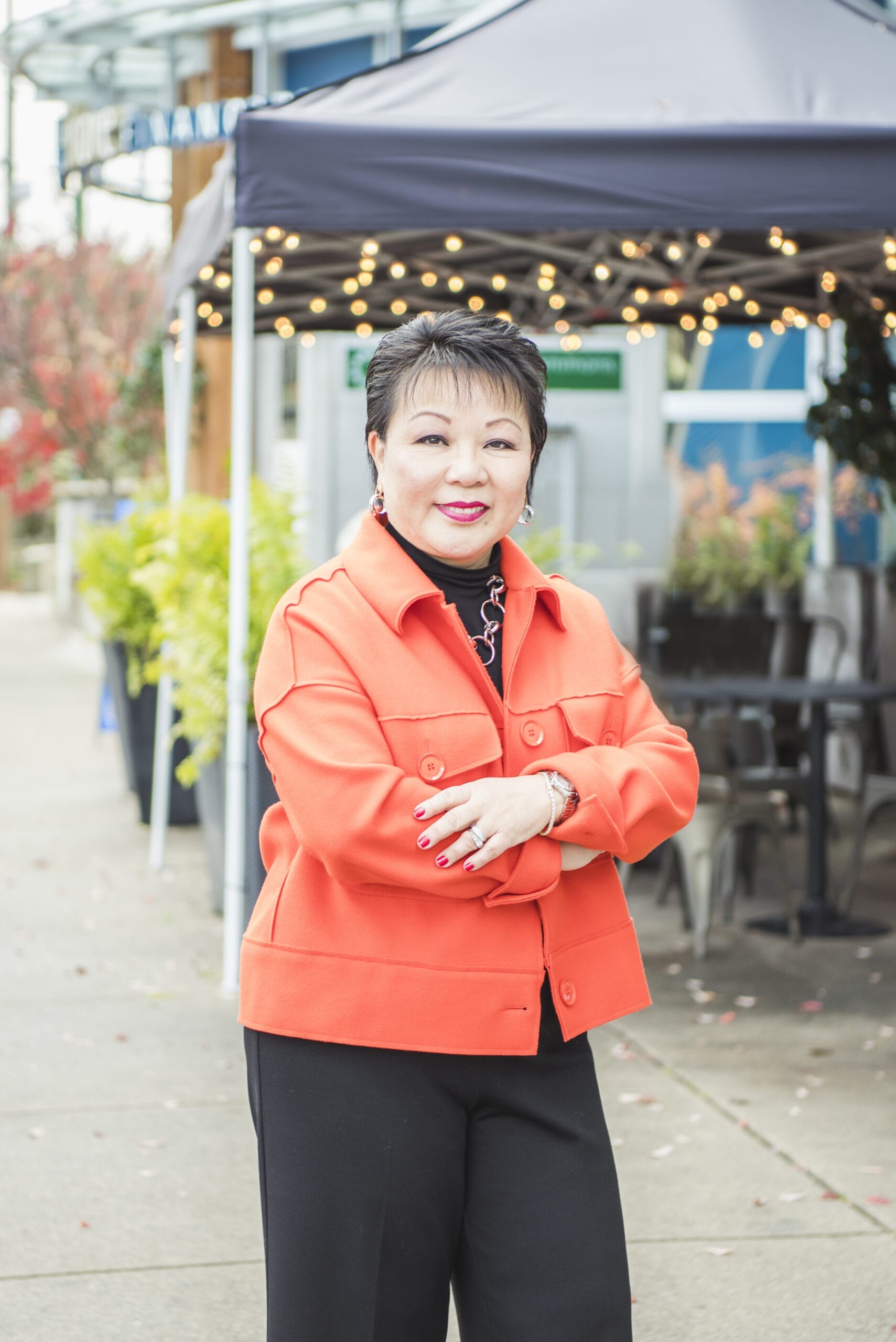 Linda Chu is in an orange jacket and stands outdoors in front of a tent with string lights.