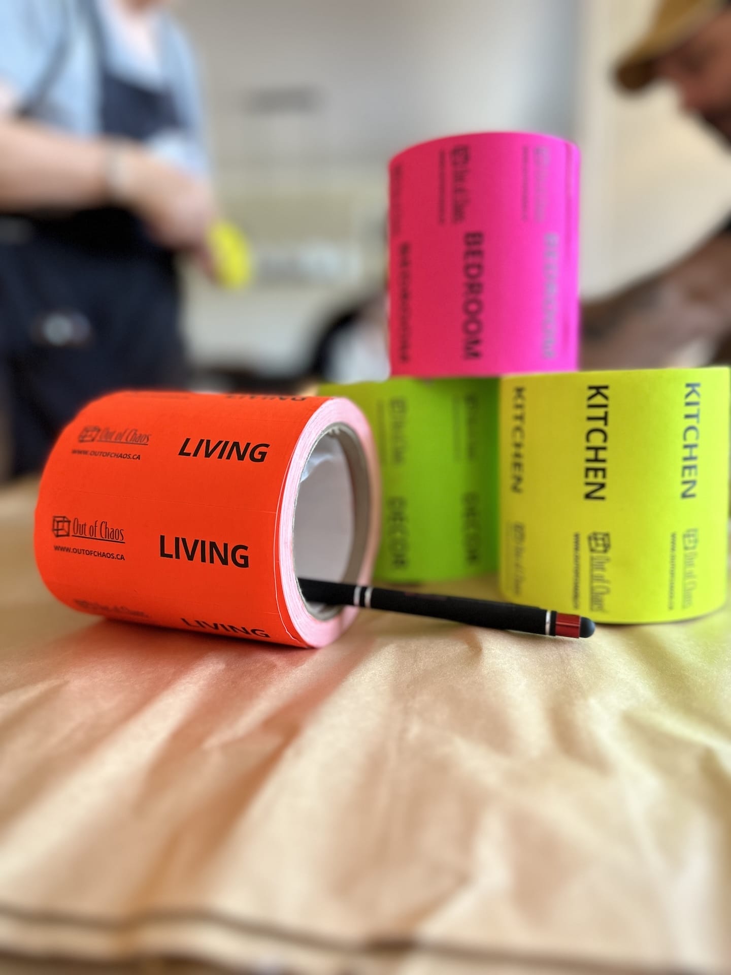 Rolls of brightly coloured moving labels for different rooms, including "Living," "Bedroom," and "Kitchen," with a black marker on a beige surface, blurred figures in the background.