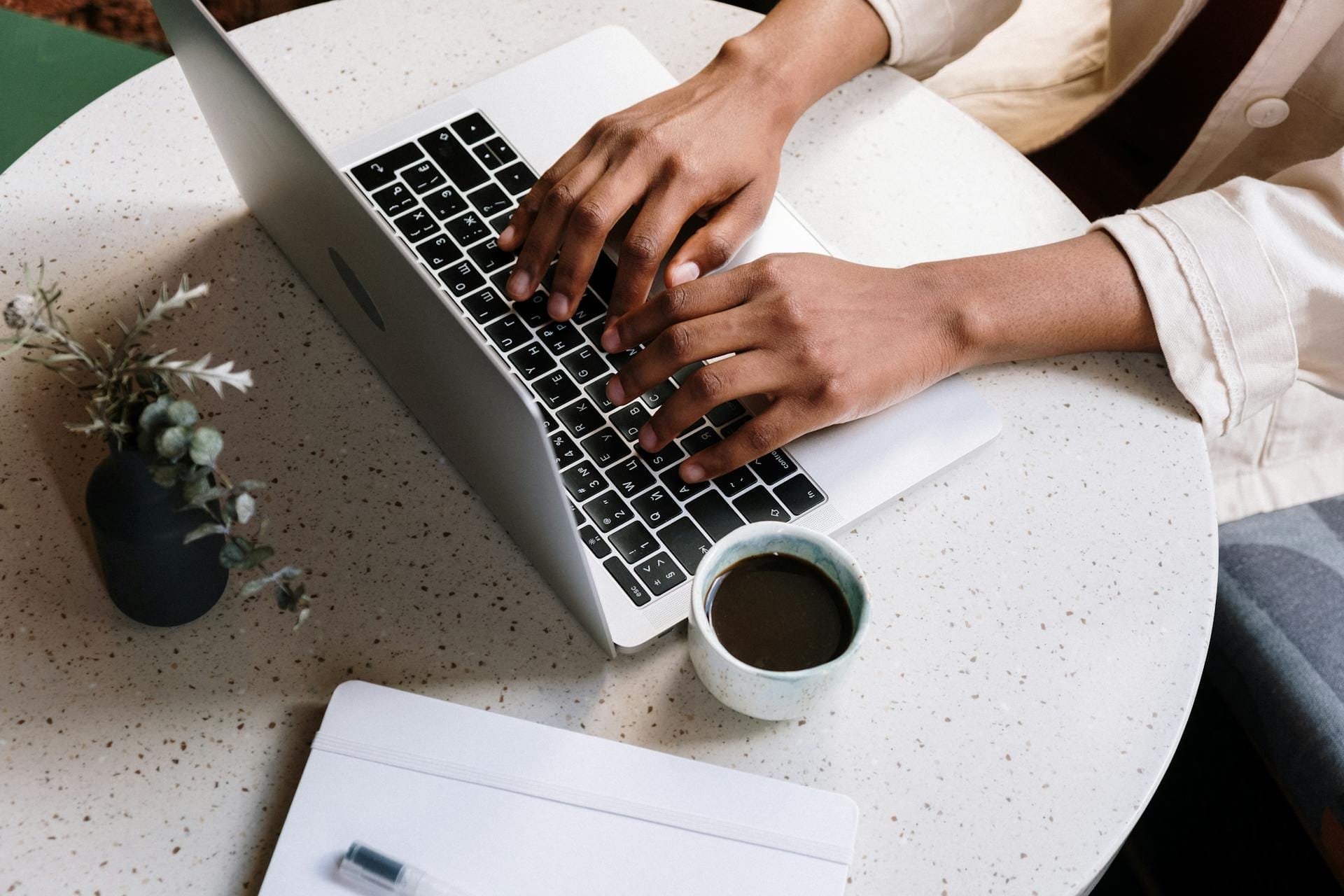 Person typing on a laptop at a round table with a cup of coffee and a closed notebook with a pen beside it.