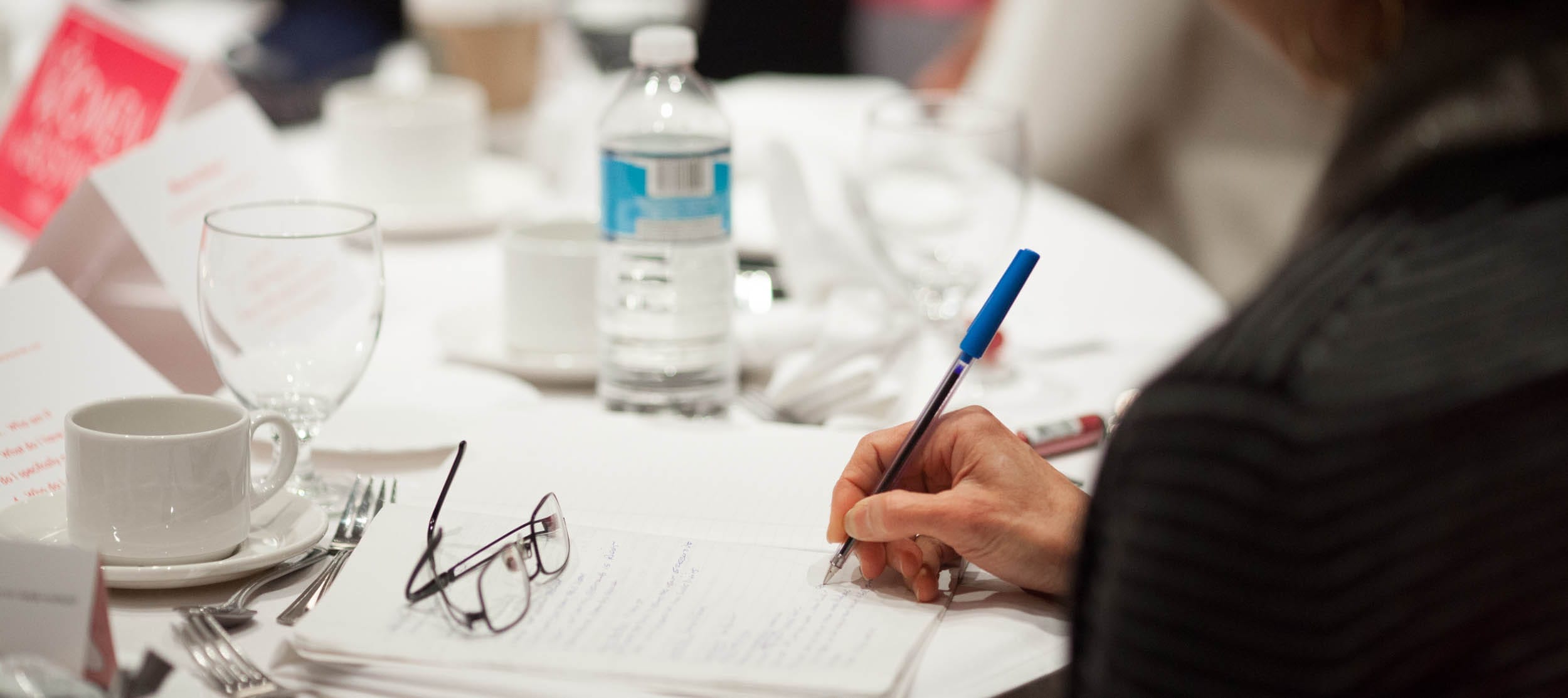 Close-up of a person writing notes with a blue pen at a table set for an event, with glasses, a water bottle, and tableware in the background