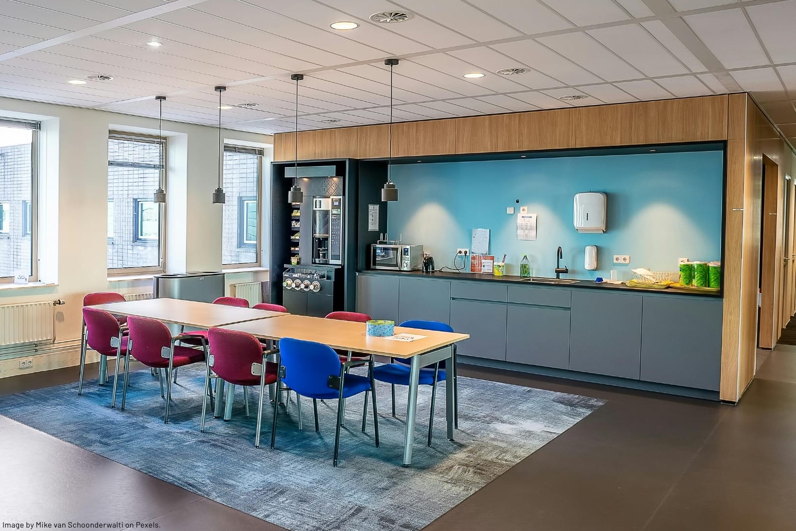 a lunchroom with a counter, cupboards, appliances along a wall with a long table and chairs in the middle of an open space is an example of a workplace common area